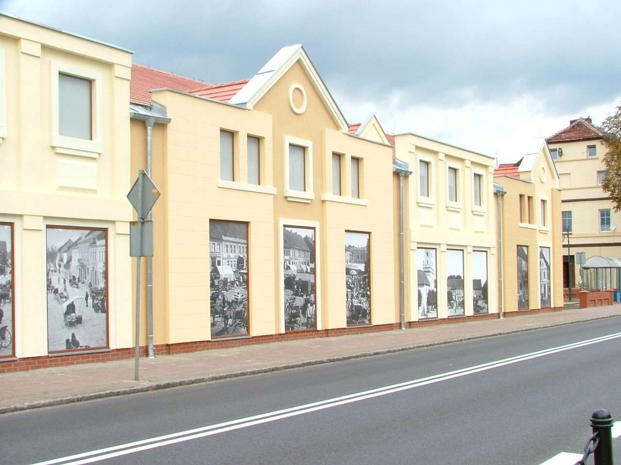 Fasada szeregu budynków pomalowanych na kremowo i żółto, z wkomponowanymi czarno-białymi fotografiami w stylu retro na parterze, widok z ulicy w pochmurny dzień.
