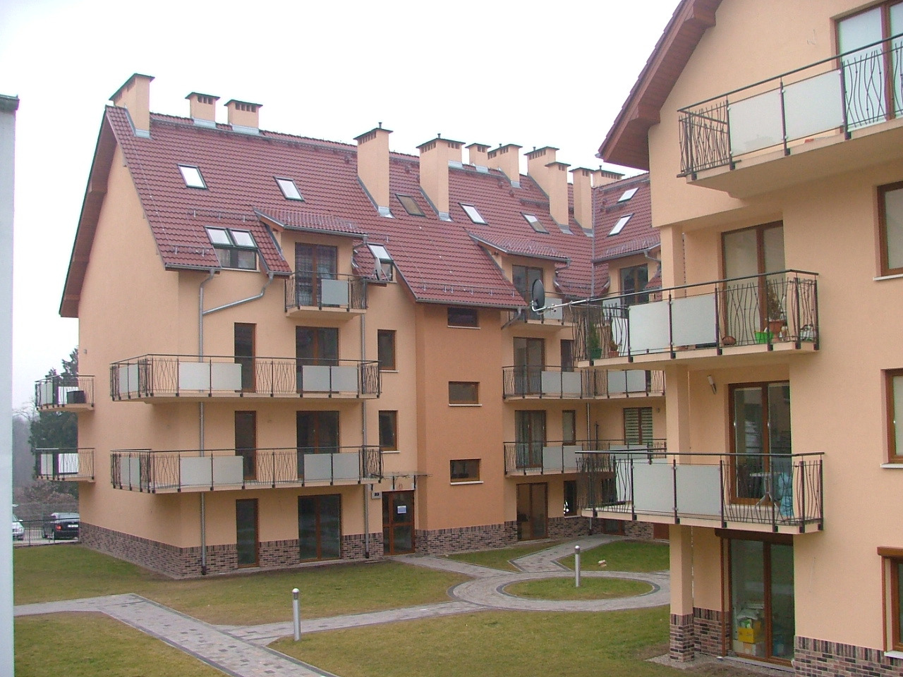 Nowoczesny budynek wielorodzinny z balkonami o jasnej elewacji i czerwonym dachem, widok z perspektywy trawnika.