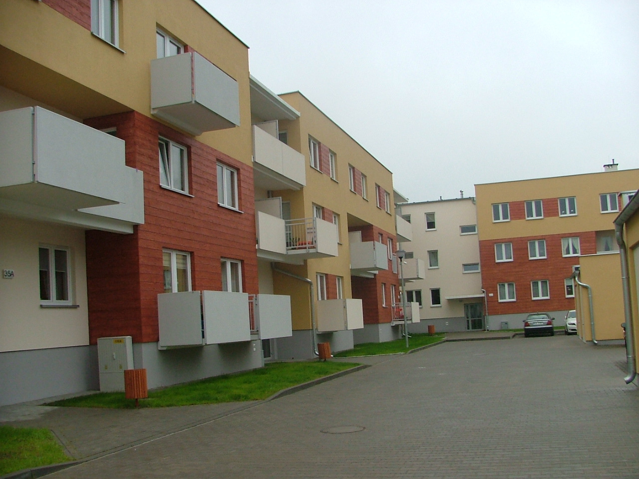 Nowoczesne osiedle mieszkaniowe z elewacją w kolorach beżu, czerwieni i bieli, balkony z białymi balustradami, widok z poziomu chodnika na dziedziniec z zaparkowanymi samochodami w pochmurny dzień.