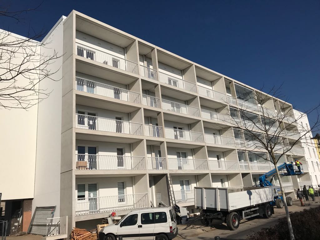 Wielopiętrowy budynek mieszkalny z balkonami, widoczna platforma robocza z pracownikiem, samochód dostawczy i materiały budowlane u podnóża budynku, błękitne niebo w tle.