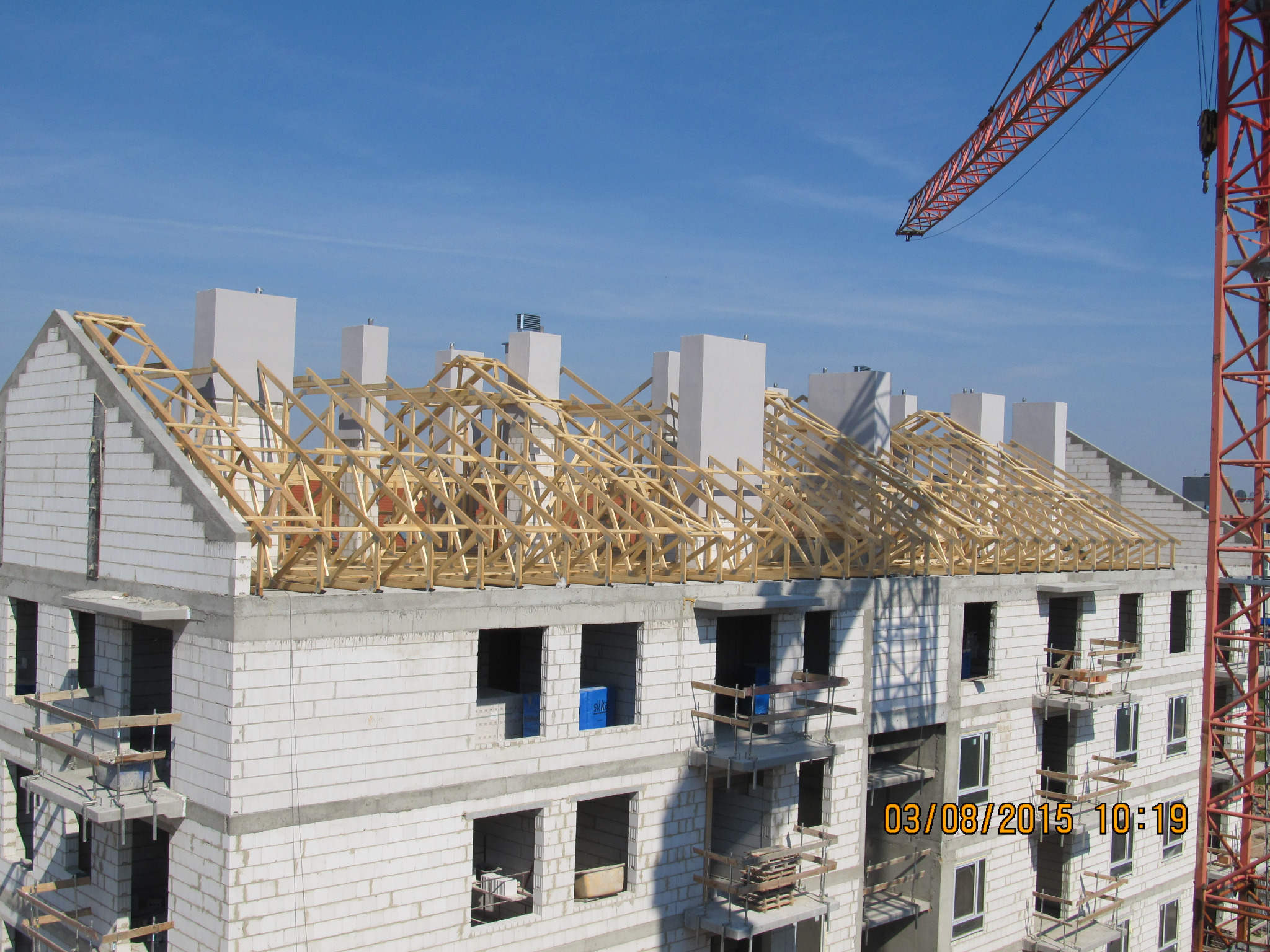 Budowa domu wielorodzinnego z białych bloczków, widoczne drewniane elementy konstrukcji dachu, kominy i fragment żurawia budowlanego na tle błękitnego nieba.