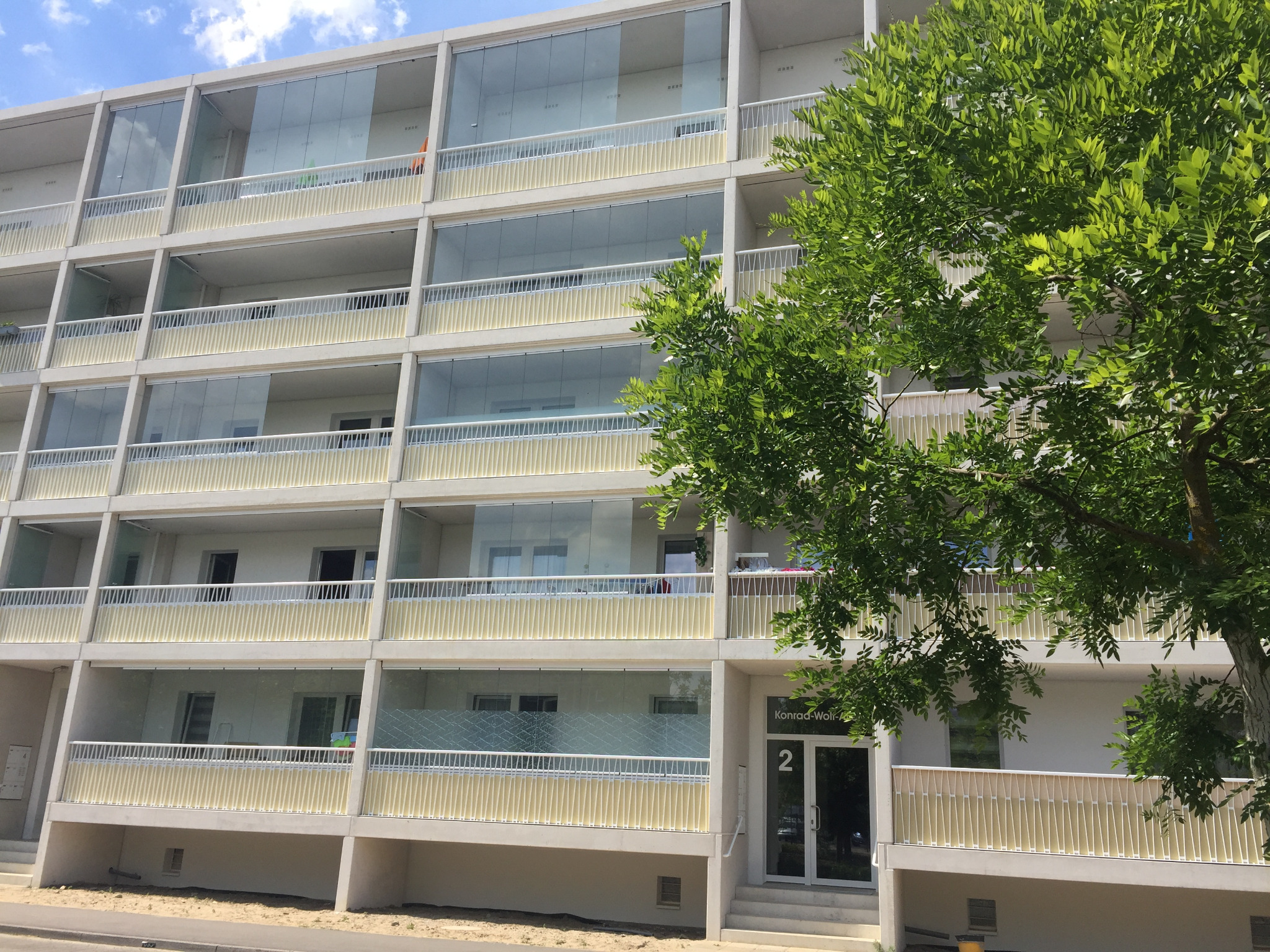 Fasada nowoczesnego budynku z balkonami zabezpieczonymi szklanymi panelami i białymi balustradami, częściowo zasłonięta przez zielone drzewo