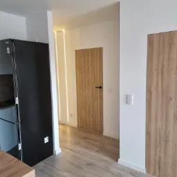 Nowoczesne wnętrze mieszkania po remoncie: panele podłogowe imitujące drewno, białe ściany z wbudowanym oświetleniem LED, drzwi wewnętrzne z okleiną drewnopodobną, czarna lodówka LG.