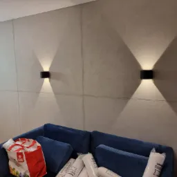 Ściana wykończona szarą gładzią imitującą beton z zamontowanymi czarnymi kinkietami, oświetlającymi przestrzeń nad granatową sofą z poduszkami i torbą z zakupami.