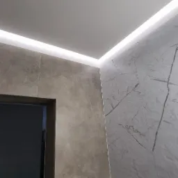 Wykończona łazienka z szarymi płytkami o zróżnicowanej fakturze, oświetleniem LED wzdłuż sufitu i widocznym fragmentem futryny drzwiowej.