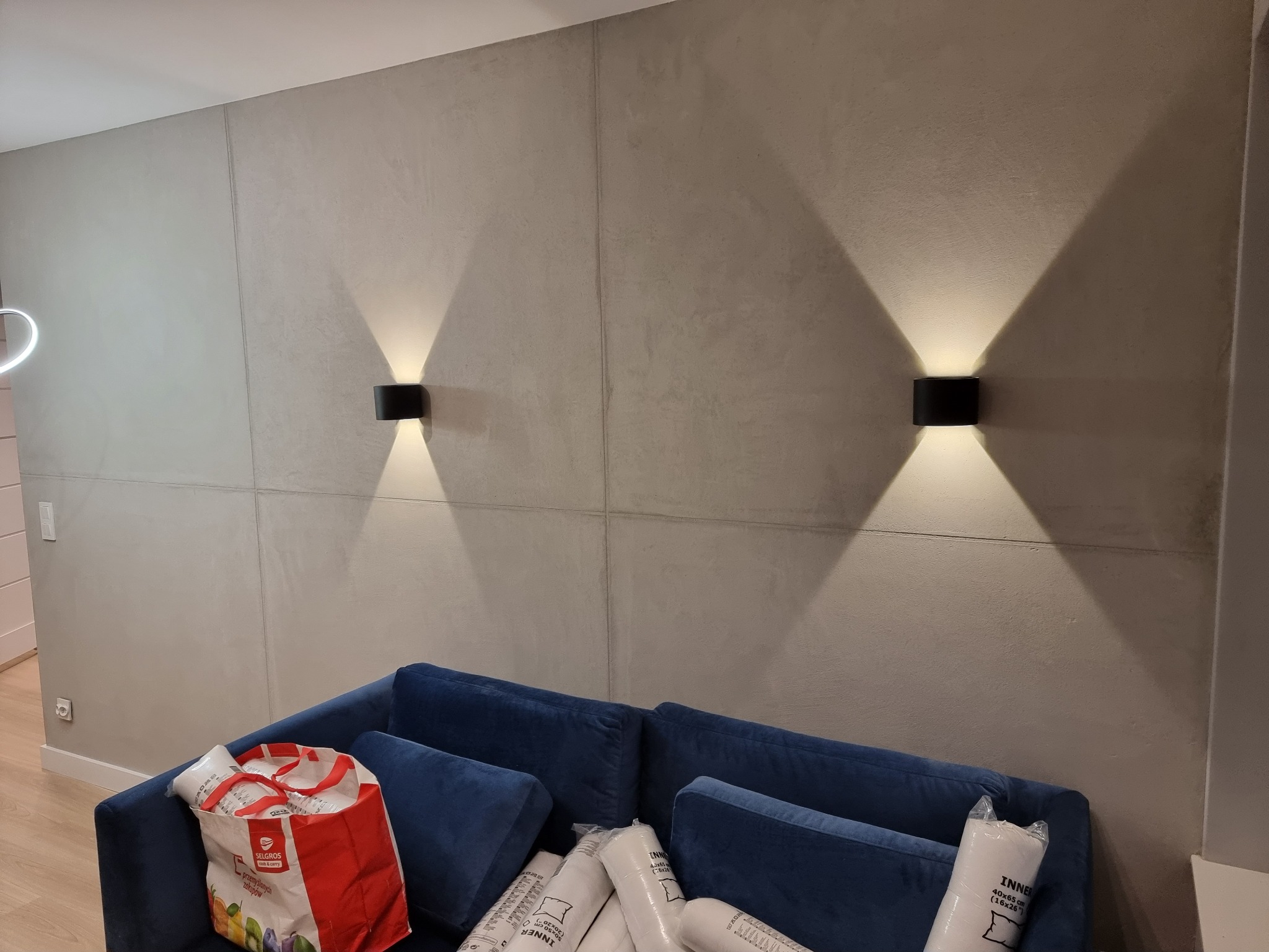 Ściana wykończona szarą gładzią imitującą beton z zamontowanymi czarnymi kinkietami, oświetlającymi przestrzeń nad granatową sofą z poduszkami i torbą z zakupami.