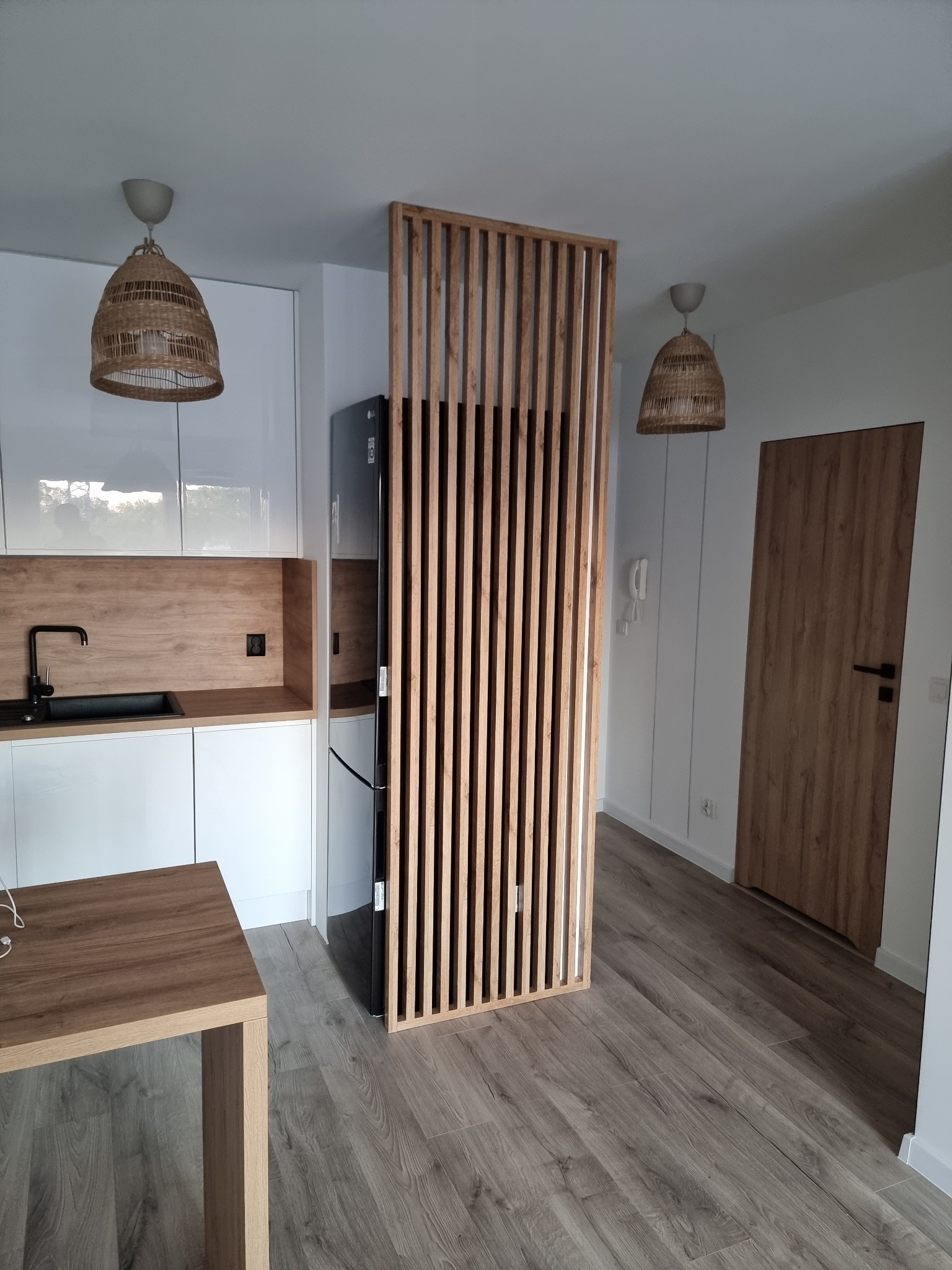 Nowoczesna aranżacja wnętrza z drewnianą ażurową ścianką działową, jasnymi meblami kuchennymi z drewnianym blatem i podłogą imitującą drewno, widoczne również lampy z abażurami w stylu boho.