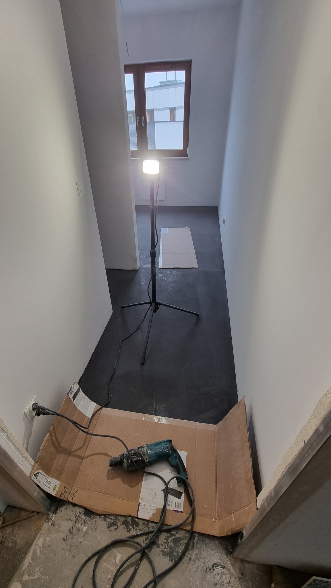 Przygotowanie do prac wykończeniowych: wiertarka udarowa na kartonie przy ścianie, lampa budowlana oświetla świeżo wylaną, czarną posadzkę w korytarzu z oknem.
