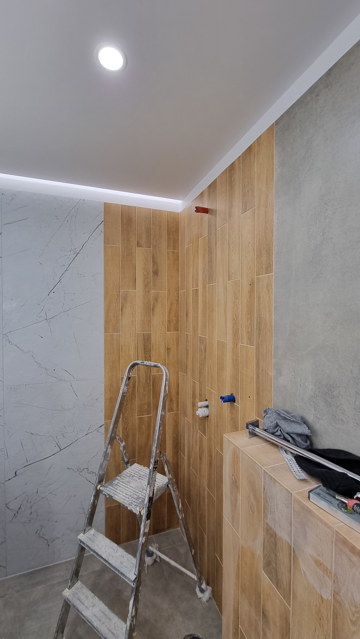 Łazienka w trakcie remontu: ściany wyłożone płytkami imitującymi drewno i marmur, aluminiowa drabina z zabrudzeniami po zaprawie, widoczne instalacje wodne, narzędzia na obudowie wanny.