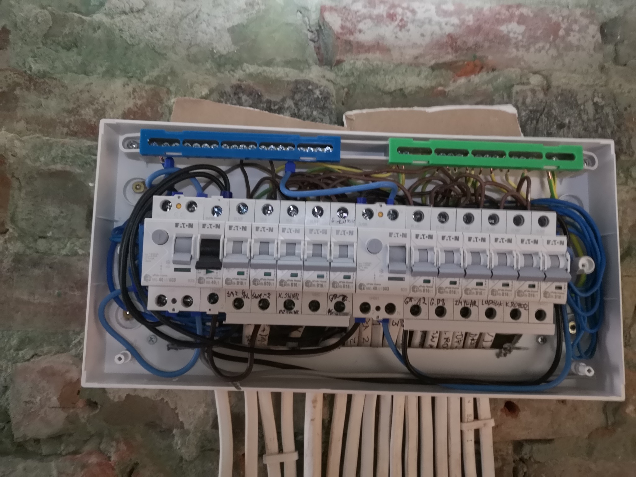 Rozdzielnica elektryczna z bezpiecznikami Eaton zamontowana na ceglanej ścianie, z widocznymi przewodami i listwami zaciskowymi w kolorach niebieskim i zielonym.