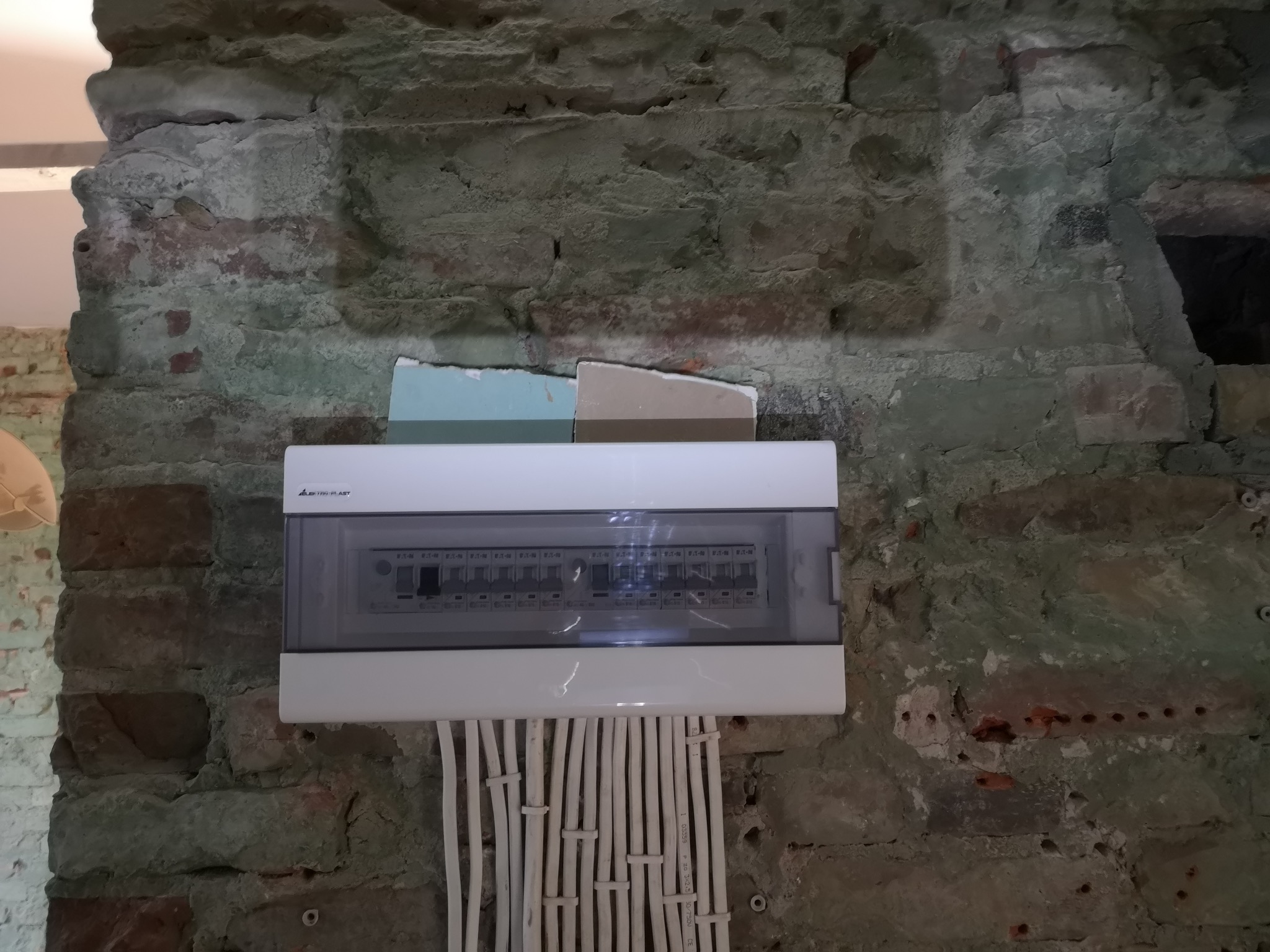 Nowa biała skrzynka rozdzielcza zamontowana na starej, surowej ścianie z cegły, z widocznymi przewodami elektrycznymi w białych peszlach.