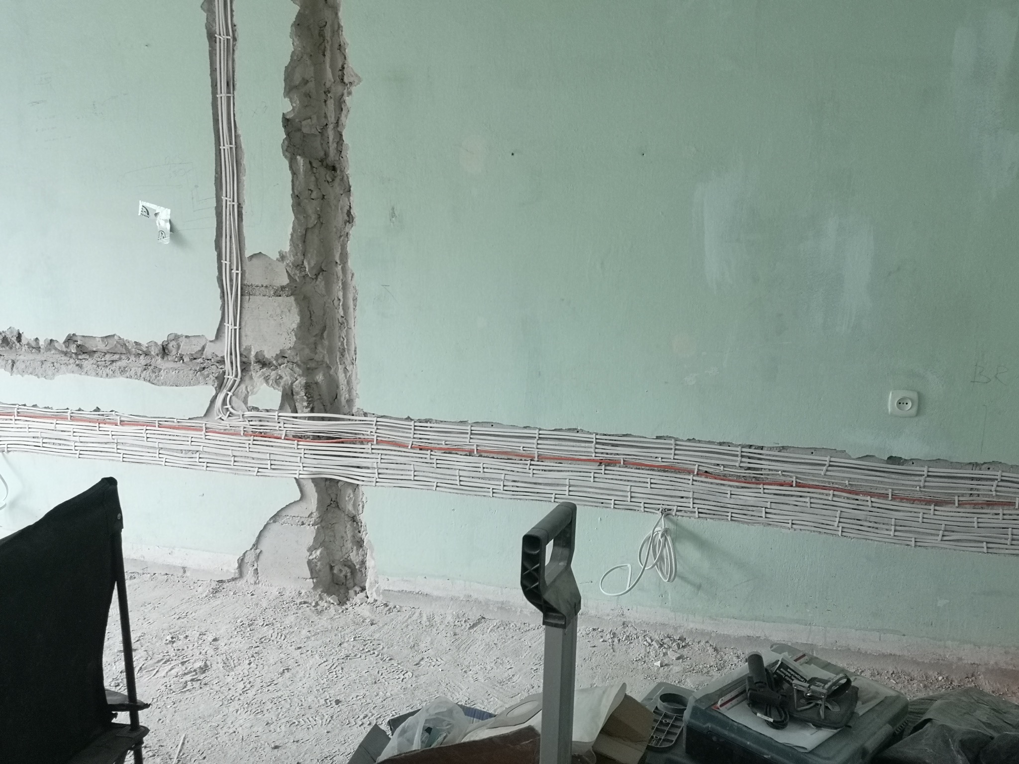 Ściana w trakcie remontu z nowo poprowadzonymi kablami elektrycznymi w korytach, widoczne ślady po kuciu w ścianie, gniazdko elektryczne na ścianie, na podłodze gruz i pył.
