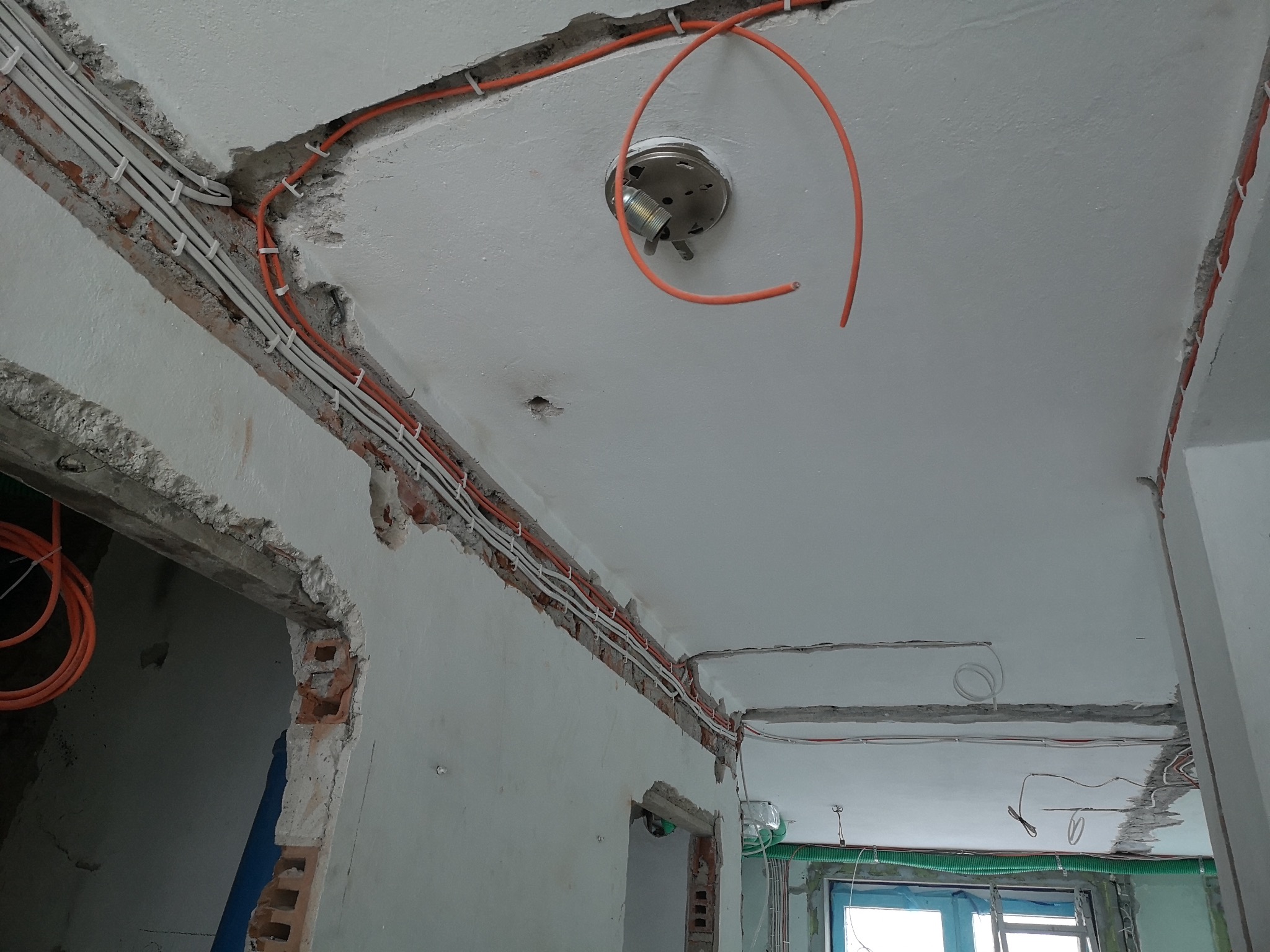 Pomieszczenie w trakcie remontu, z widocznymi przewodami elektrycznymi prowadzonymi w bruzdach w ścianach i suficie, oraz puszką pod lampę sufitową z wystającym przewodem.