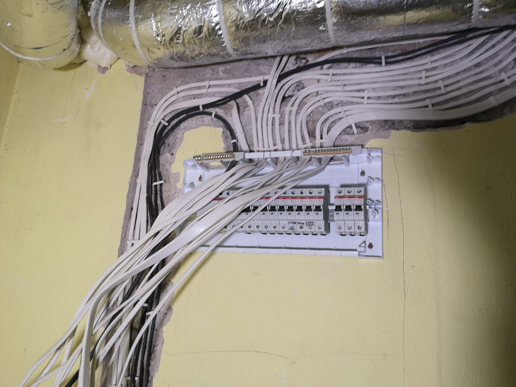 Rozdzielnica elektryczna z licznymi białymi i czarnymi przewodami ułożonymi w łuk, zamontowana na żółtej ścianie w pobliżu rury wentylacyjnej.