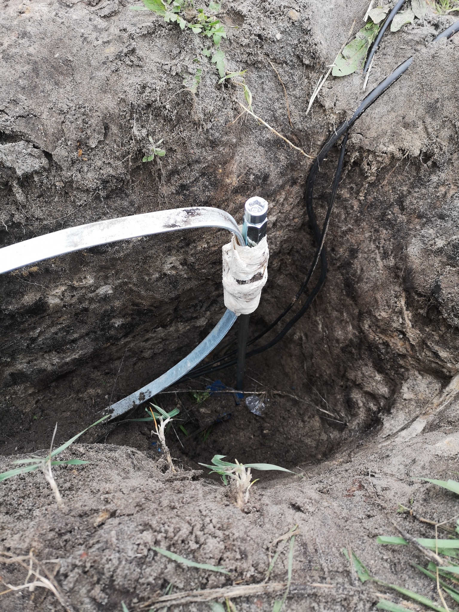 Uziemienie w wykopie, przewody elektryczne prowadzone w ziemi, element metalowy owinięty białą taśmą.