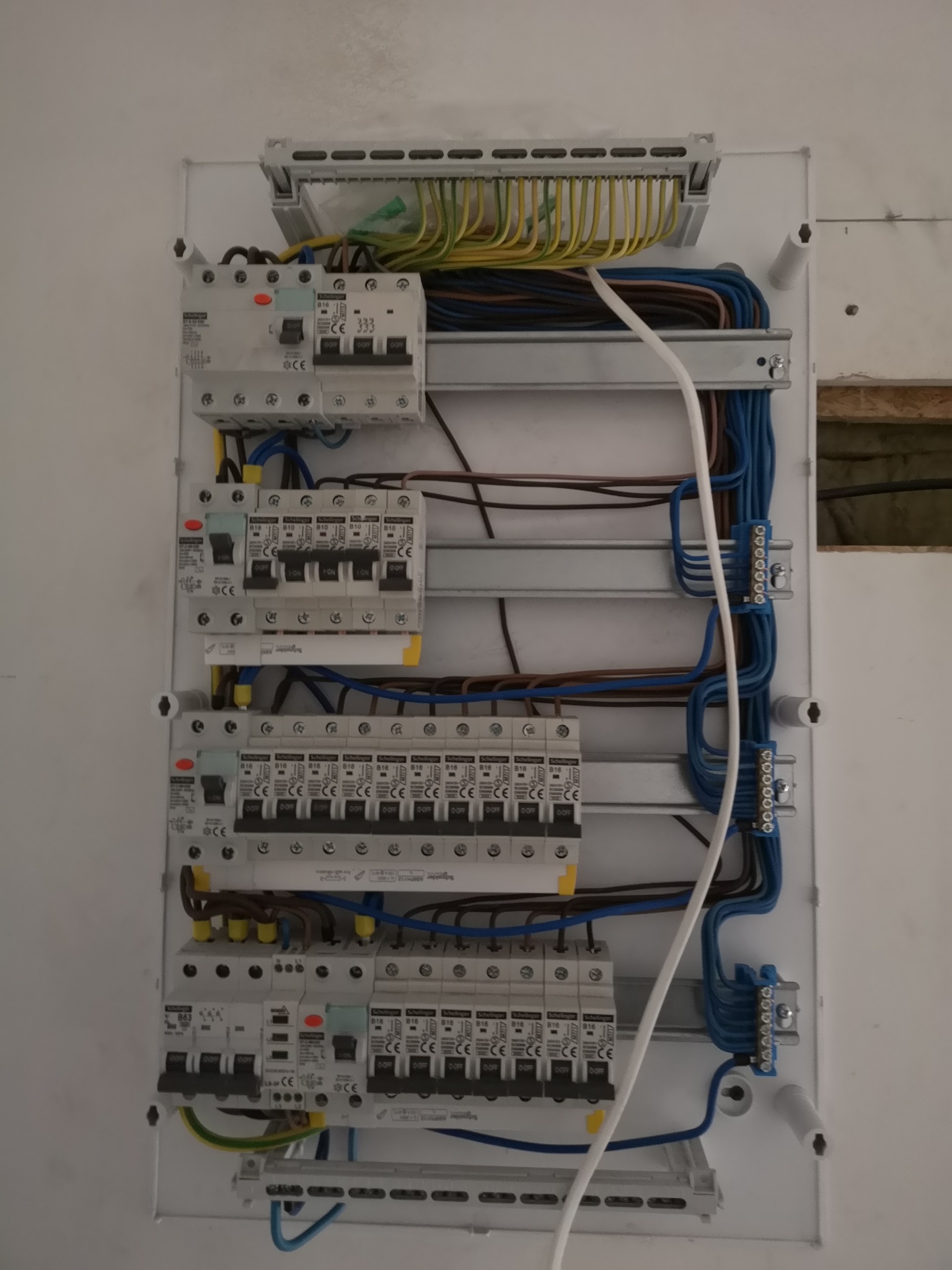 Rozdzielnica elektryczna z zainstalowanymi wyłącznikami różnicowoprądowymi i bezpiecznikami, widoczne przewody w kolorach niebieskim, brązowym, żółto-zielonym, uporządkowane w listwach. Częściowo...