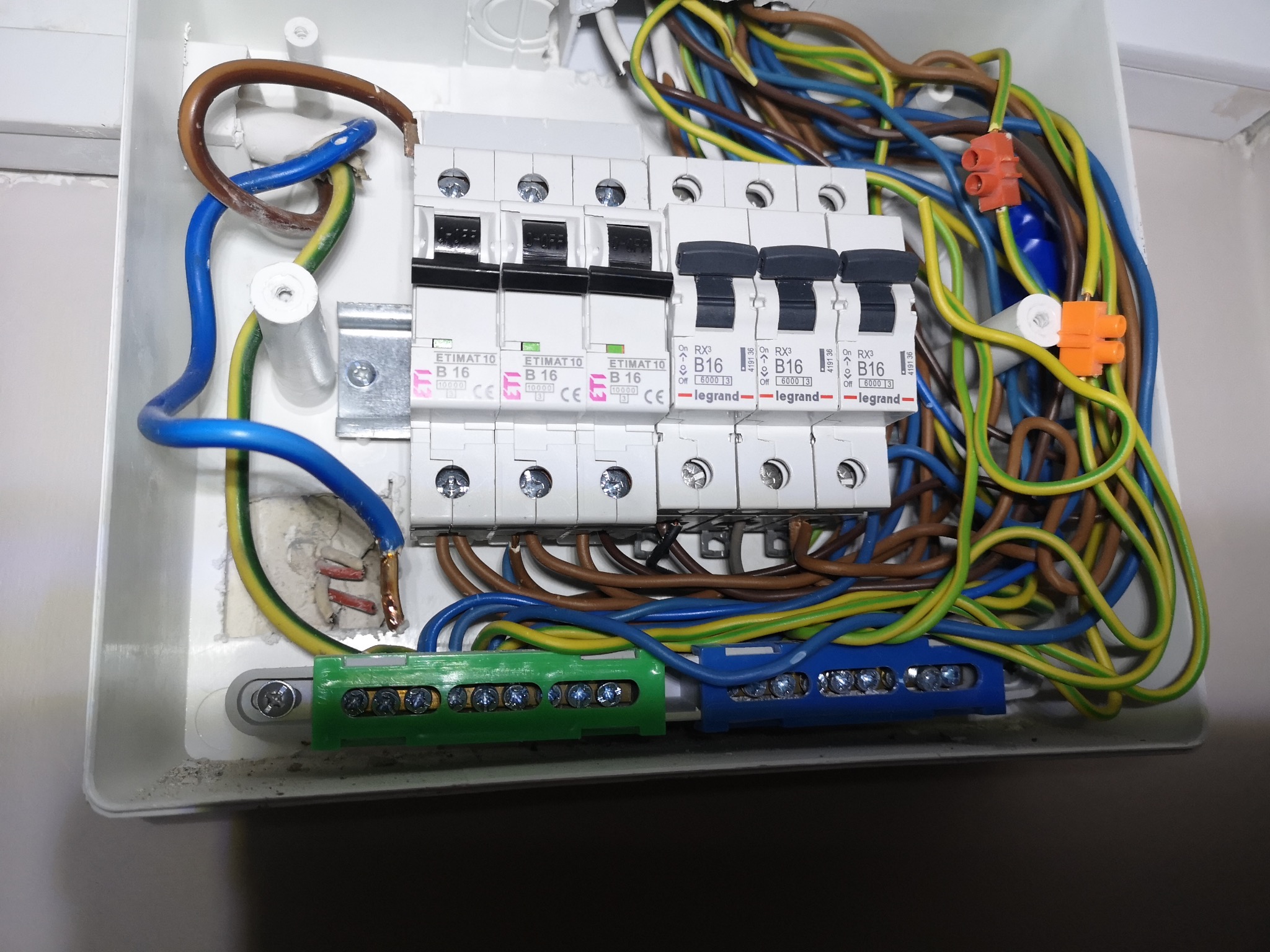 Otwarta skrzynka elektryczna z widocznymi bezpiecznikami, przewodami w różnych kolorach (niebieski, brązowy, żółto-zielony) oraz złączkami instalacyjnymi. Widoczne ślady montażu na ścianie.