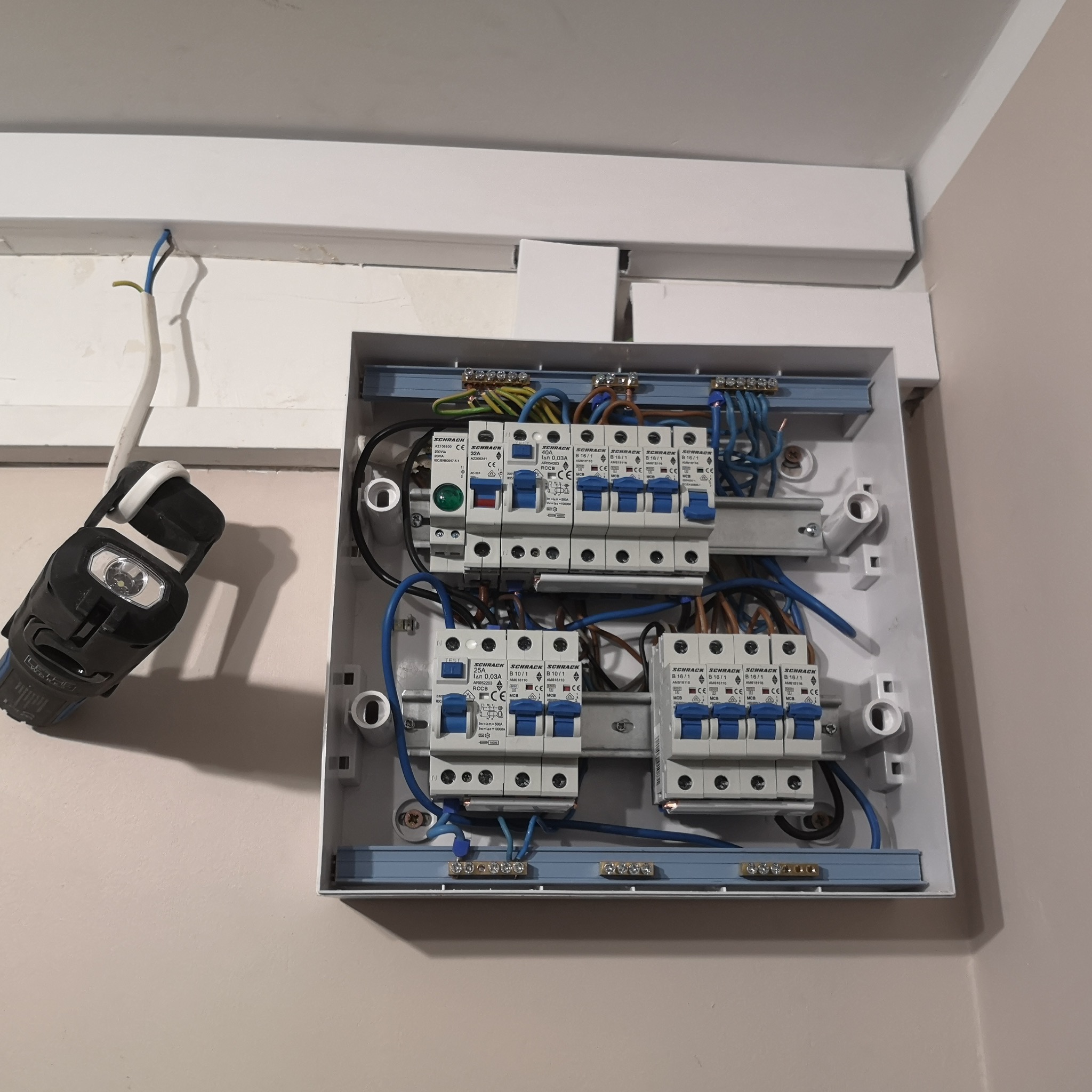 Rozdzielnica elektryczna z zamontowanymi bezpiecznikami i okablowaniem, oświetlona przenośną lampą LED, widok z bliska.