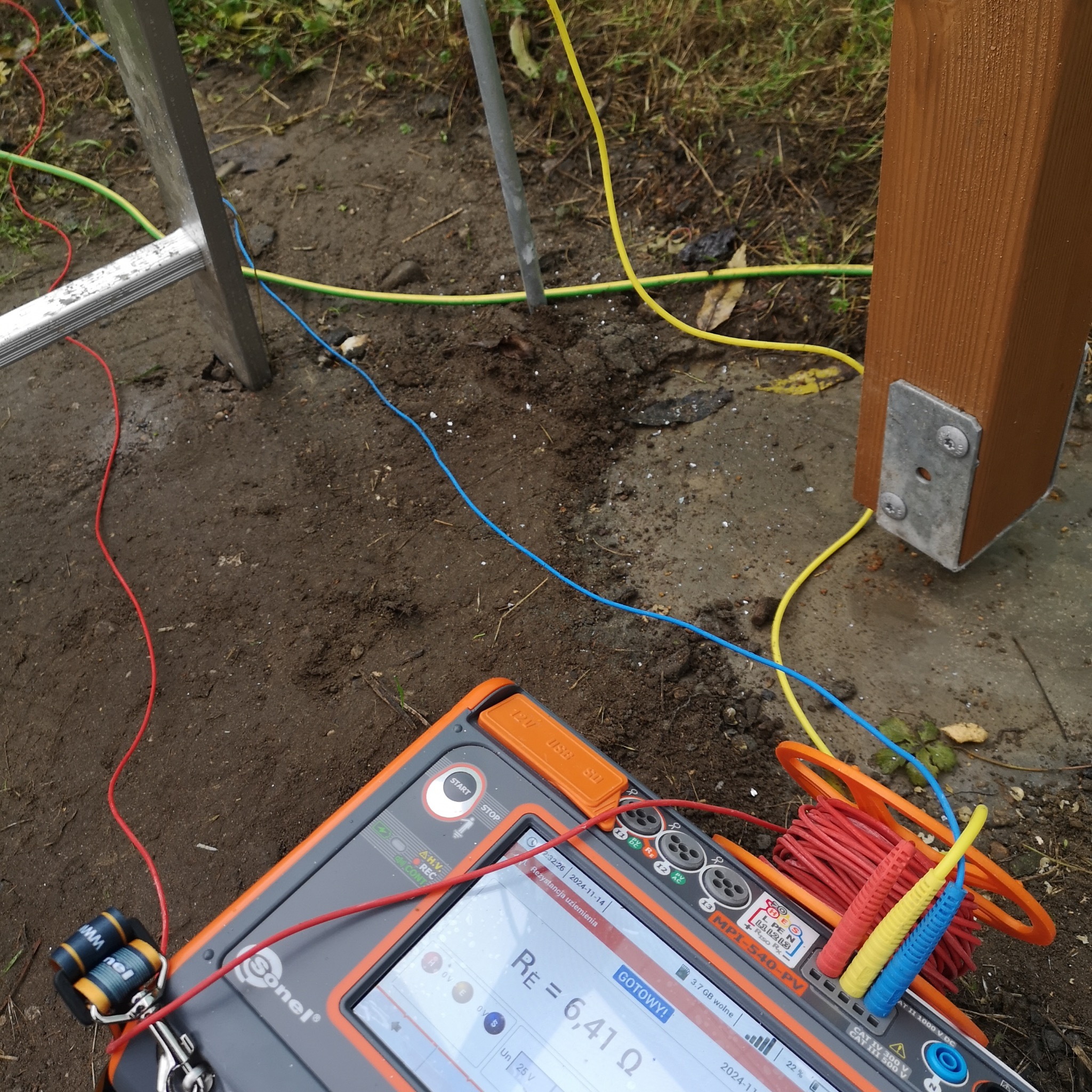 Pomiar rezystancji uziemienia za pomocą miernika Sonel MPI-540-PV, widoczne przewody pomiarowe w kolorach czerwonym, niebieskim i żółtym podłączone do uziomu w gruncie obok drewnianego słupka.