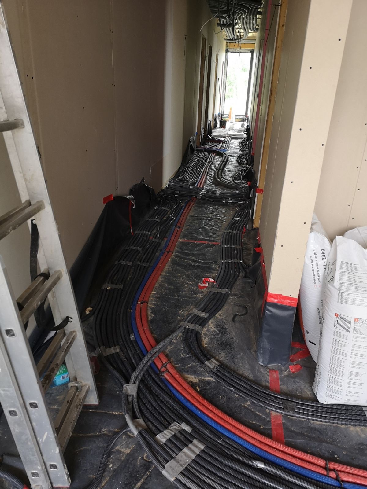 Wnętrze w trakcie prac: liczne kable elektryczne i rury prowadzone pod podłogą, zabezpieczone folią, widoczna drabina i worki z materiałami budowlanymi w tle.