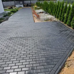 Nowo ułożona, szara kostka brukowa prowadząca do altany w ogrodzie, obrzeża wykończone ciemniejszą kostką, w tle rząd tuj.