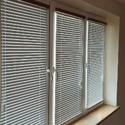 Trzy okna z białymi żaluzjami poziomymi, drewniane wykończenie u góry, parapet z jasnego drewna, ściana w tle z ciemnozieloną tapetą w kwiaty i ptaki.