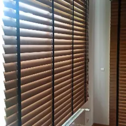 Drewniane żaluzje z ciemnymi taśmami na oknie, obok biały grzejnik, parkiet na podłodze.