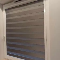 Okno z zamontowaną roletą dzień-noc w odcieniach szarości, widoczny mechanizm sterujący i biała rama okienna.