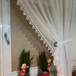 Przejrzysta, biała firanka z koronkowym wykończeniem zawieszona przy oknie, dekorowana dwoma małymi choinkami w czerwonych doniczkach z pluszowymi misiami w strojach świątecznych.