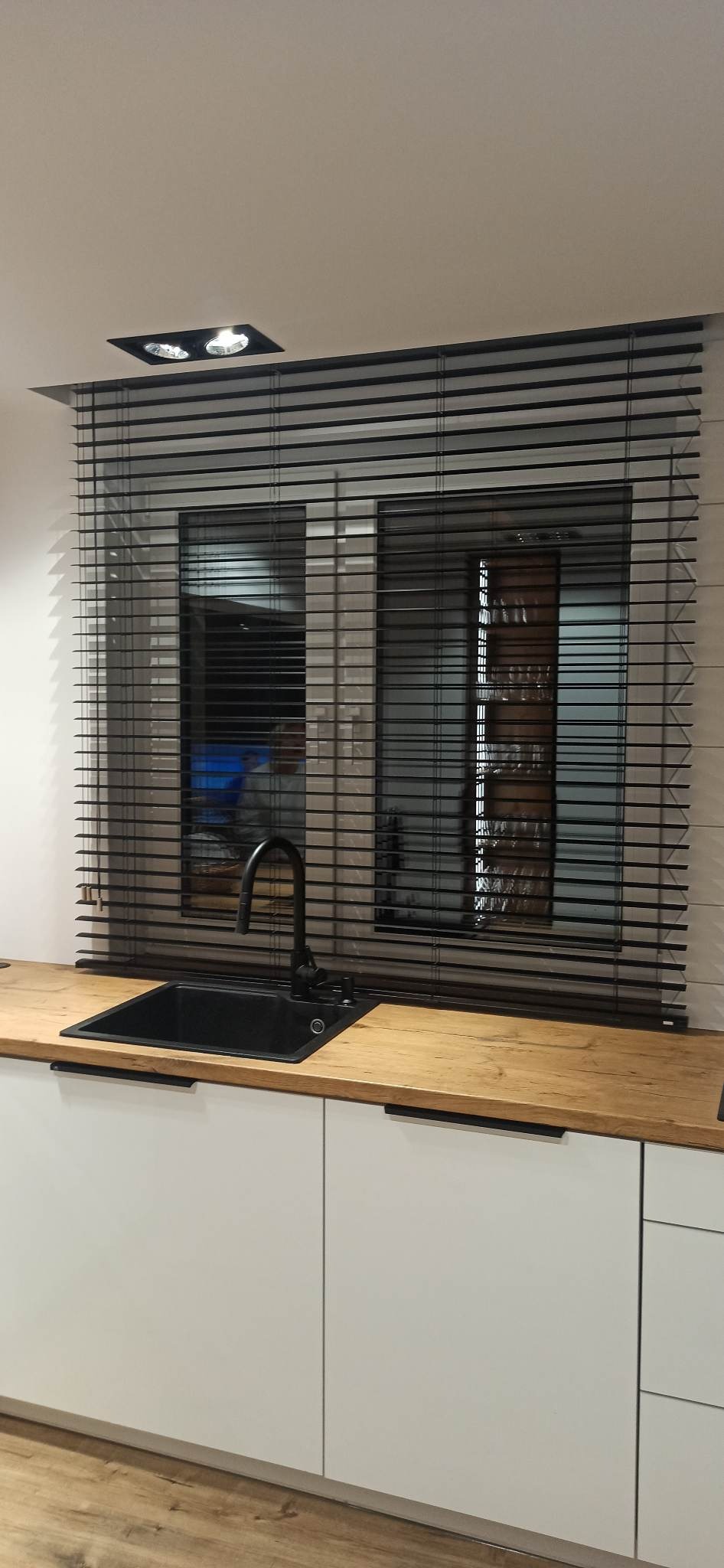 Czarne poziome listwy w oknach nad zlewem w nowoczesnej kuchni z drewnianym blatem i białymi szafkami.