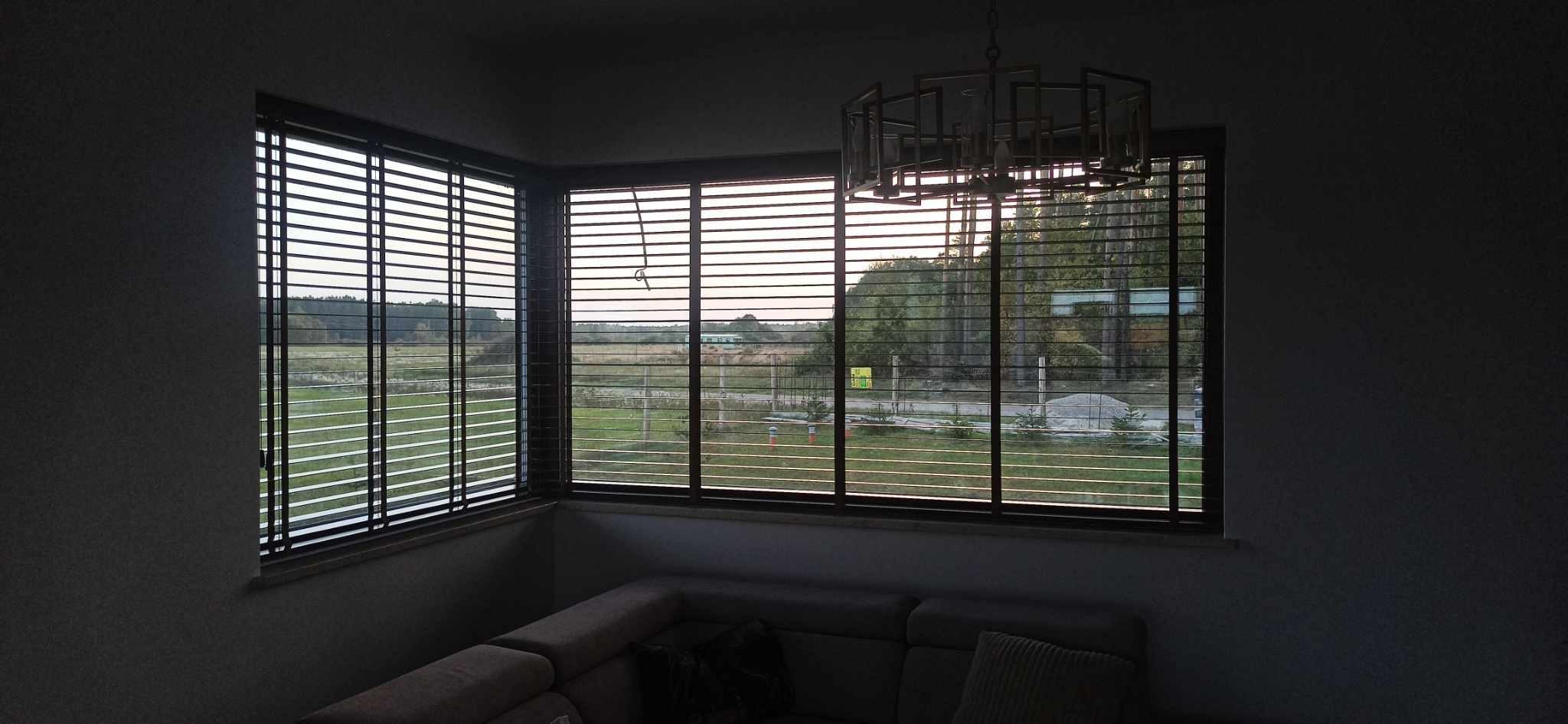 Drewniane żaluzje poziome w ciemnej ramie okiennej, widok na krajobraz za oknem, nowoczesny żyrandol w tle.