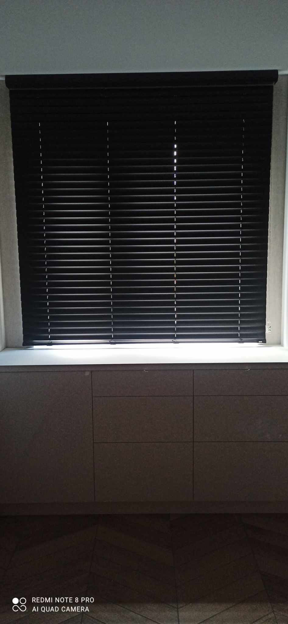 Ciemne żaluzje poziome zamontowane w oknie nad szarą zabudową meblową z szufladami.