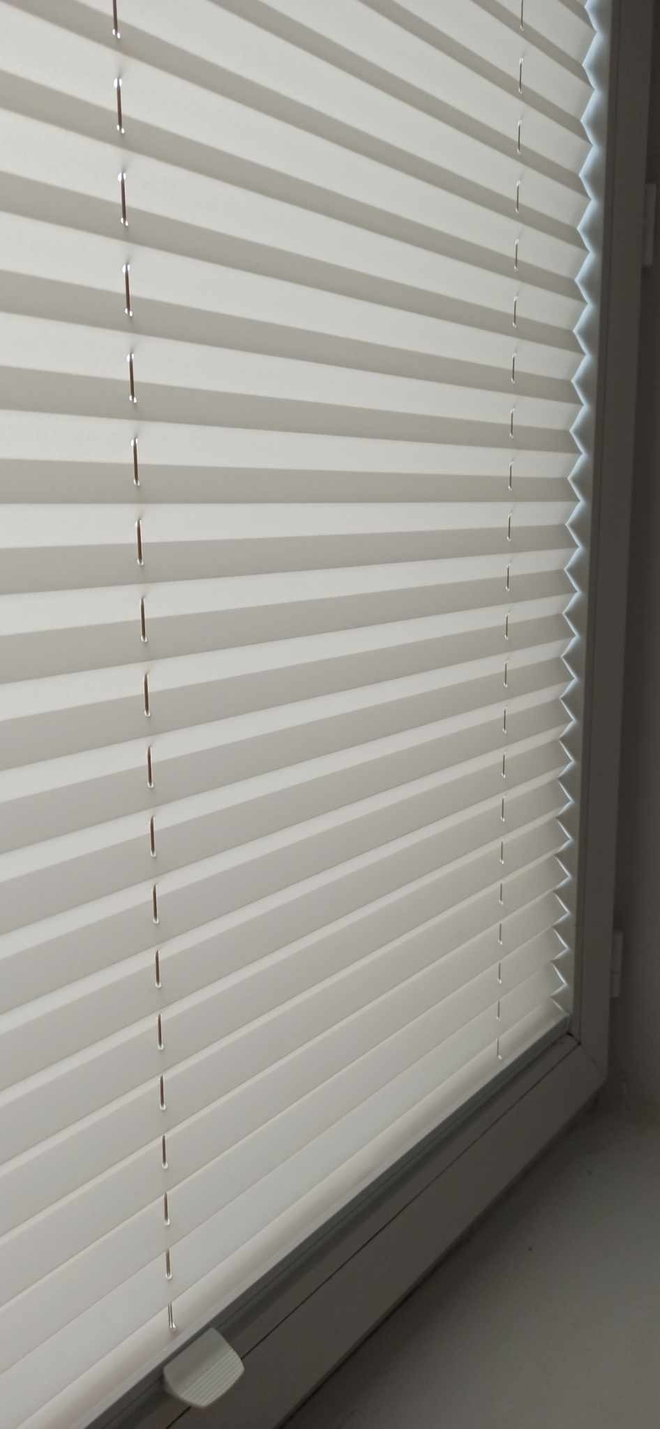 Zamknięte, białe, plisowane osłony okienne z widocznym mechanizmem sterującym, zamontowane w oknie z szarą ramą.