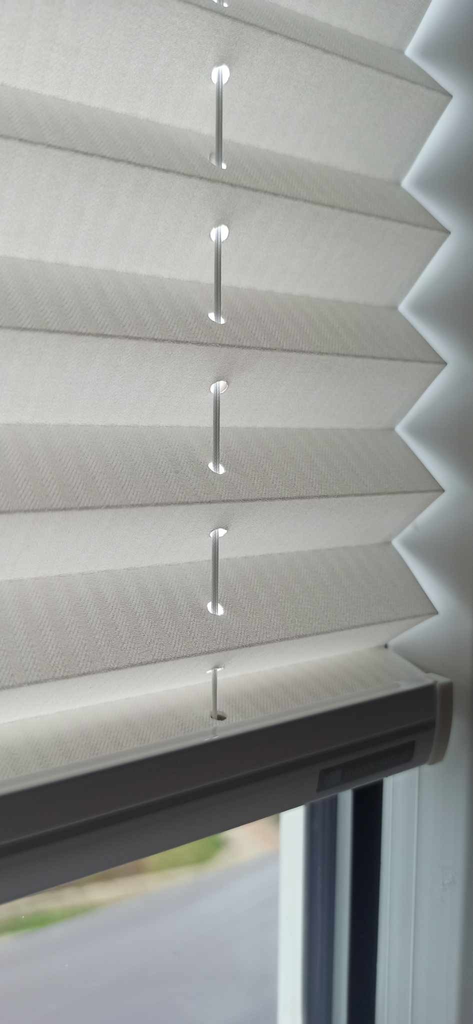 Detaliczny widok białych plis okiennych z widocznymi sznurkami i strukturą materiału, w tle widok z okna.