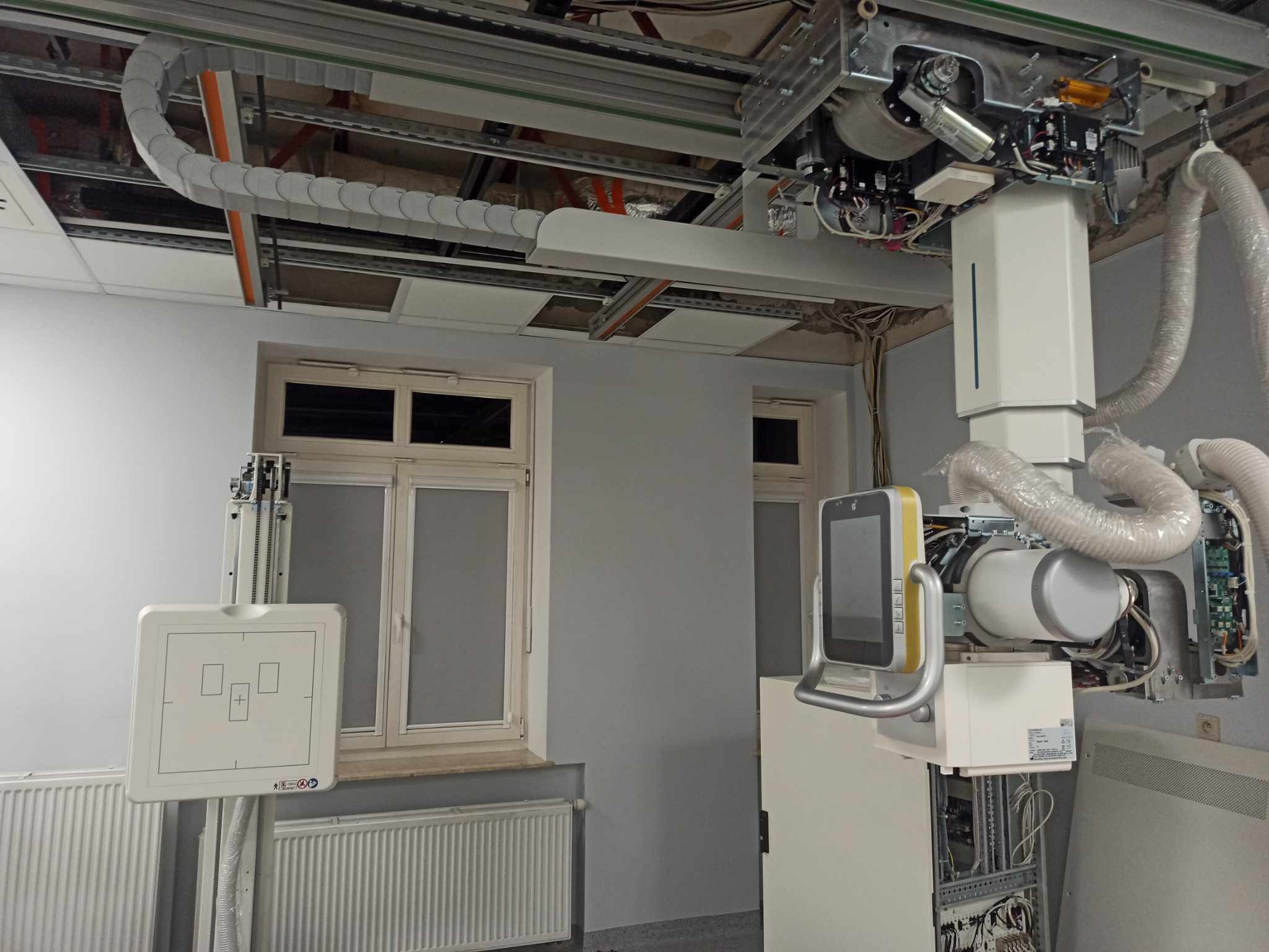 Wnętrze pomieszczenia z zainstalowanym, zaawansowanym sprzętem medycznym do diagnostyki obrazowej, widoczne kable i instalacje pod sufitem, okna w tle.