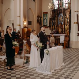 Szymon Korta Fotografia - Para młoda w kościele podczas ceremonii ślubnej, z gośćmi w tle i dekoracjami kwiatowymi. Ujęcie z perspektywy lekko z góry, ukazujące wzór posadzki.