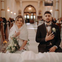 Szymon Korta Fotografia - Para młoda siedzi w kościele podczas ceremonii ślubnej, z gośćmi w tle. Panna młoda trzyma bukiet, a pan młody ma butonierkę. Ciepłe oświetlenie podkreśla uroczysty nastrój.