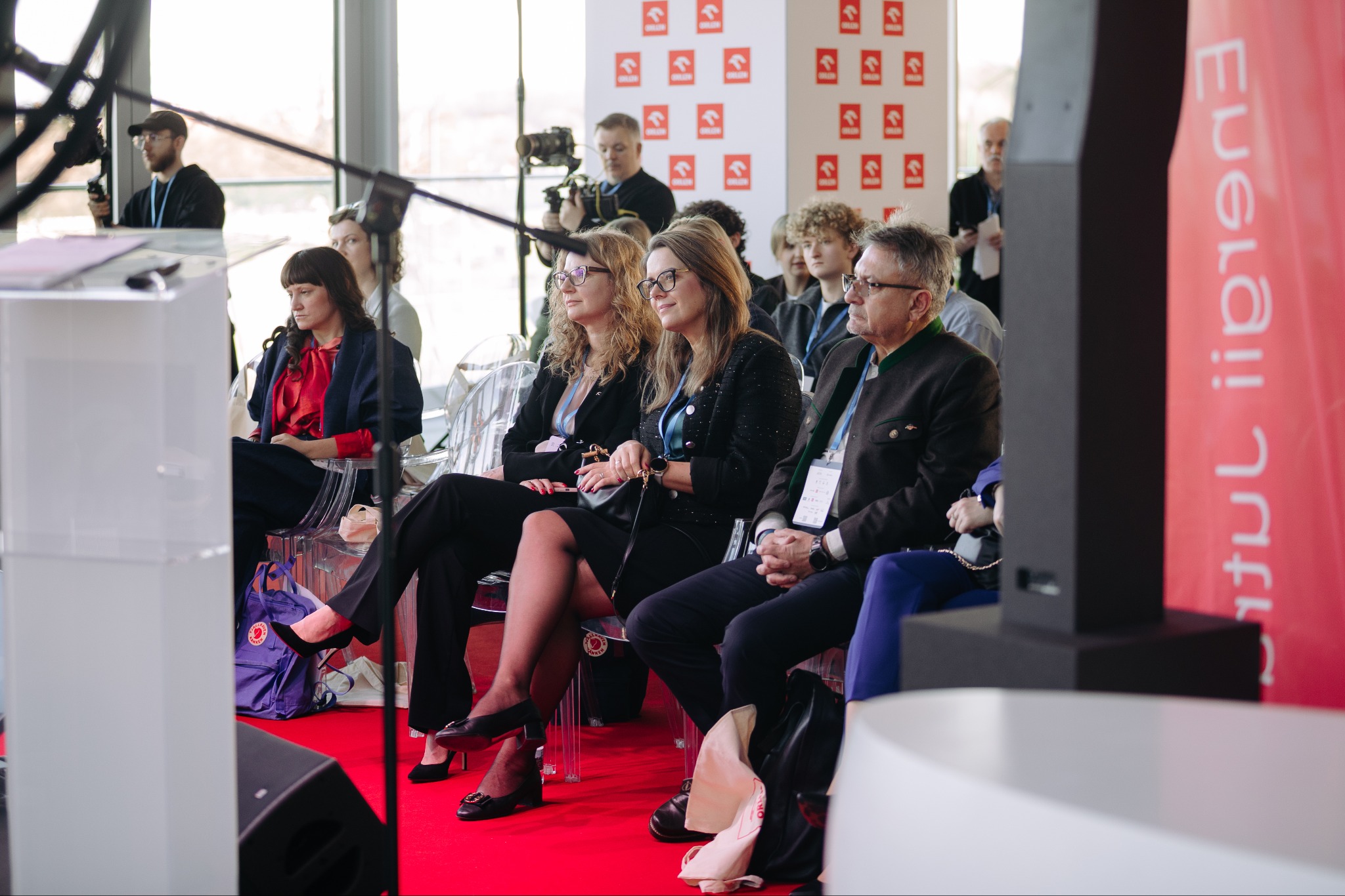 Uczestnicy konferencji siedzą na przezroczystych krzesłach, przysłuchując się prelekcji. W tle widoczni fotografowie i logo sponsora.