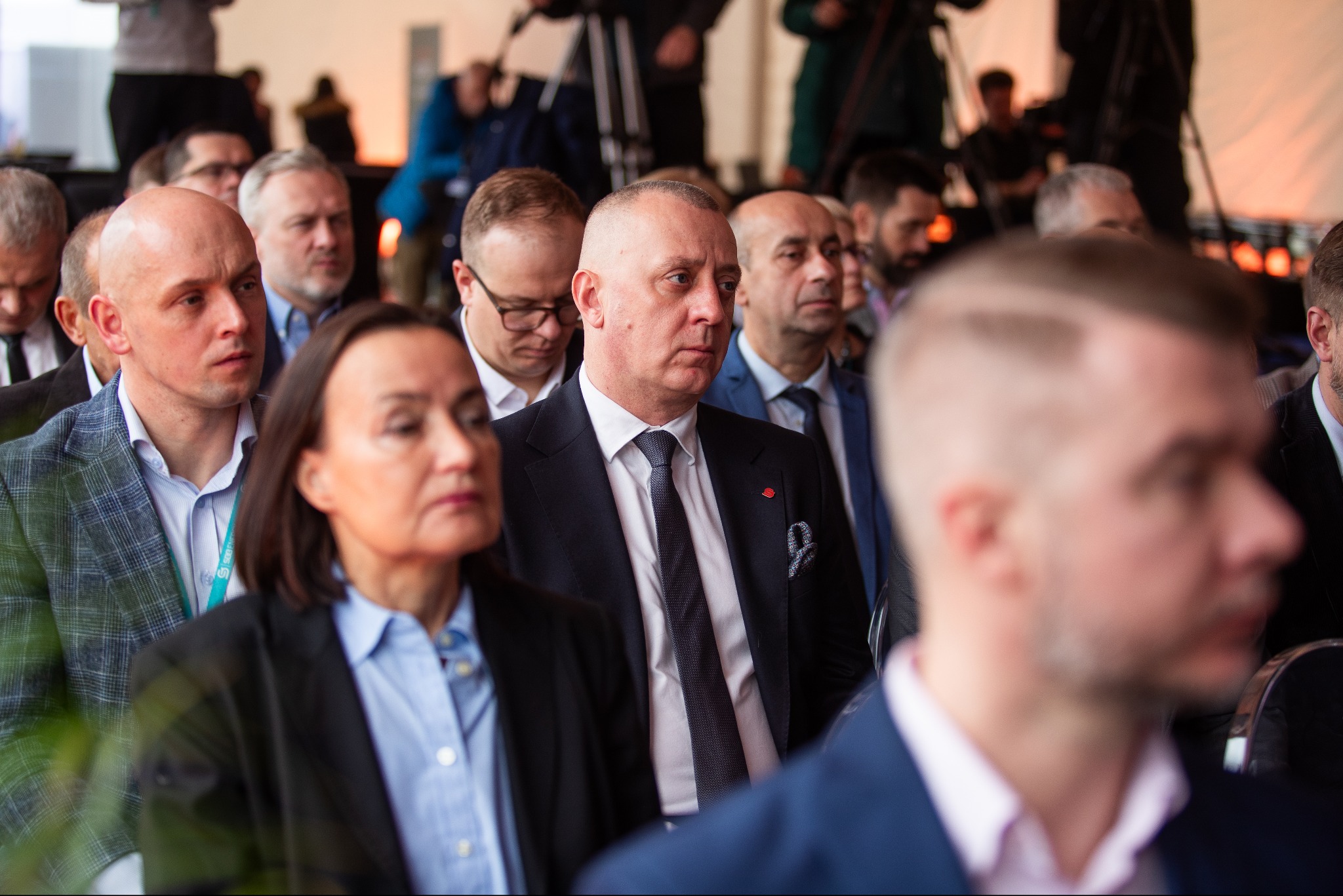 Uczestnicy konferencji PGE siedzą w rzędach, skupieni na prelegencie. Mężczyzna w garniturze z poszetką w centrum kadru. Widoczne kamery i oświetlenie sceniczne w tle.