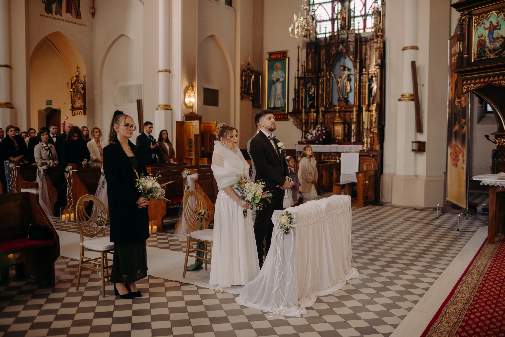 Para młoda w kościele podczas ceremonii ślubnej, z gośćmi w tle i dekoracjami kwiatowymi. Ujęcie z perspektywy lekko z góry, ukazujące wzór posadzki.
