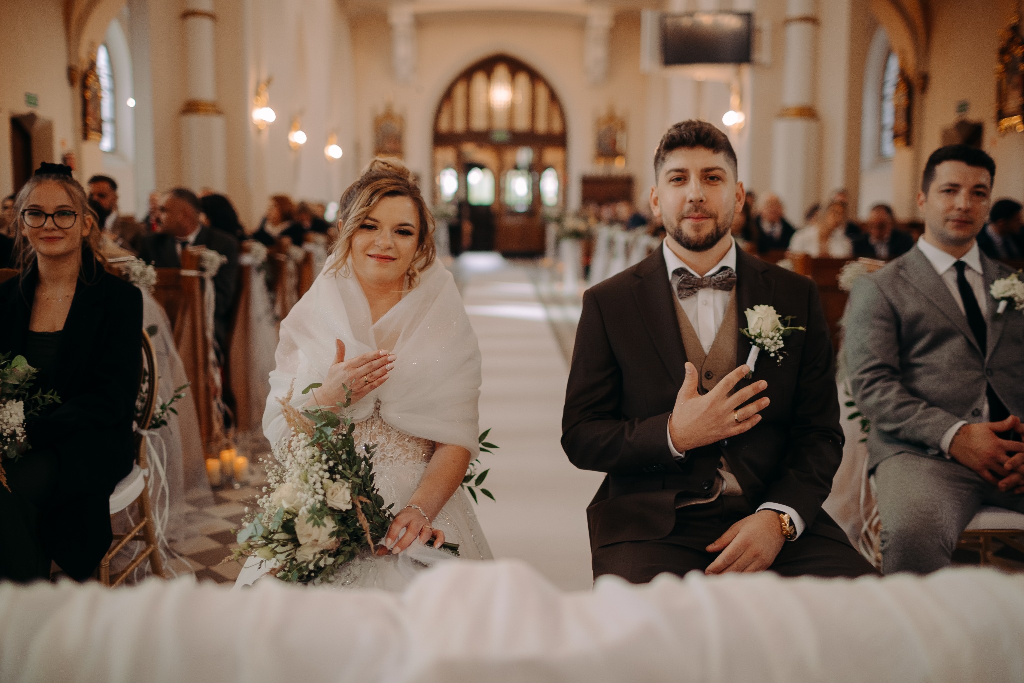 Para młoda siedzi w kościele podczas ceremonii ślubnej, z gośćmi w tle. Panna młoda trzyma bukiet, a pan młody ma butonierkę. Ciepłe oświetlenie podkreśla uroczysty nastrój.