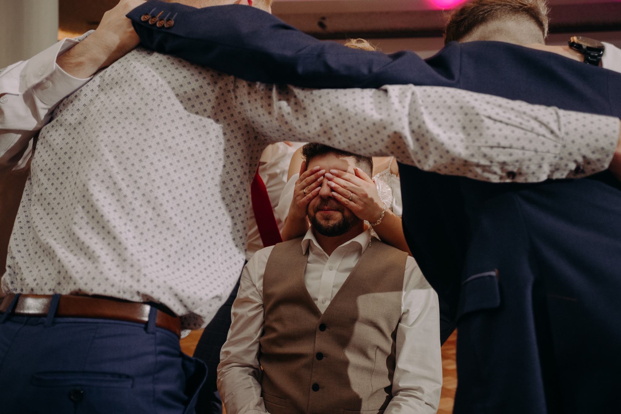 Pan młody z zasłoniętymi oczami przez pannę młodą, otoczony przez gości w garniturach podczas weselnej zabawy.