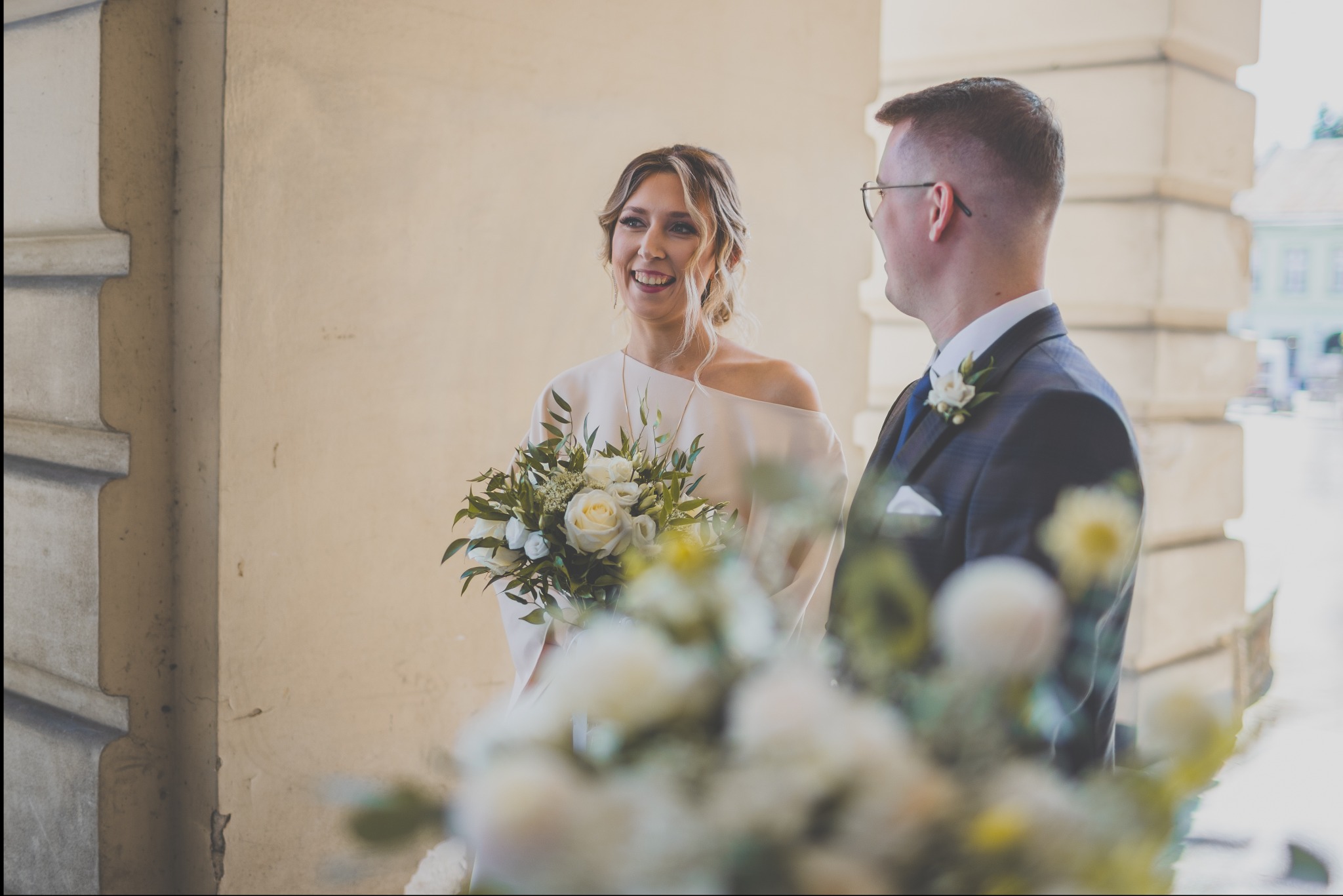 Uśmiechnięta panna młoda w białej sukni z bukietem białych róż, obok pan młody w ciemnym garniturze z butonierką, w tle kolumnada.