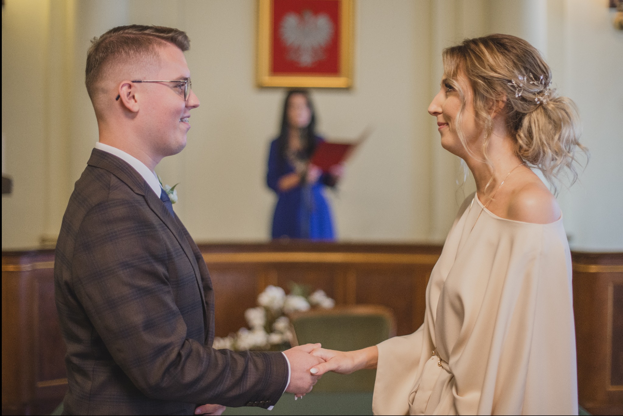 Para młoda trzymająca się za ręce podczas ceremonii ślubnej w urzędzie, w tle urzędniczka z czerwoną księgą i godło Polski.