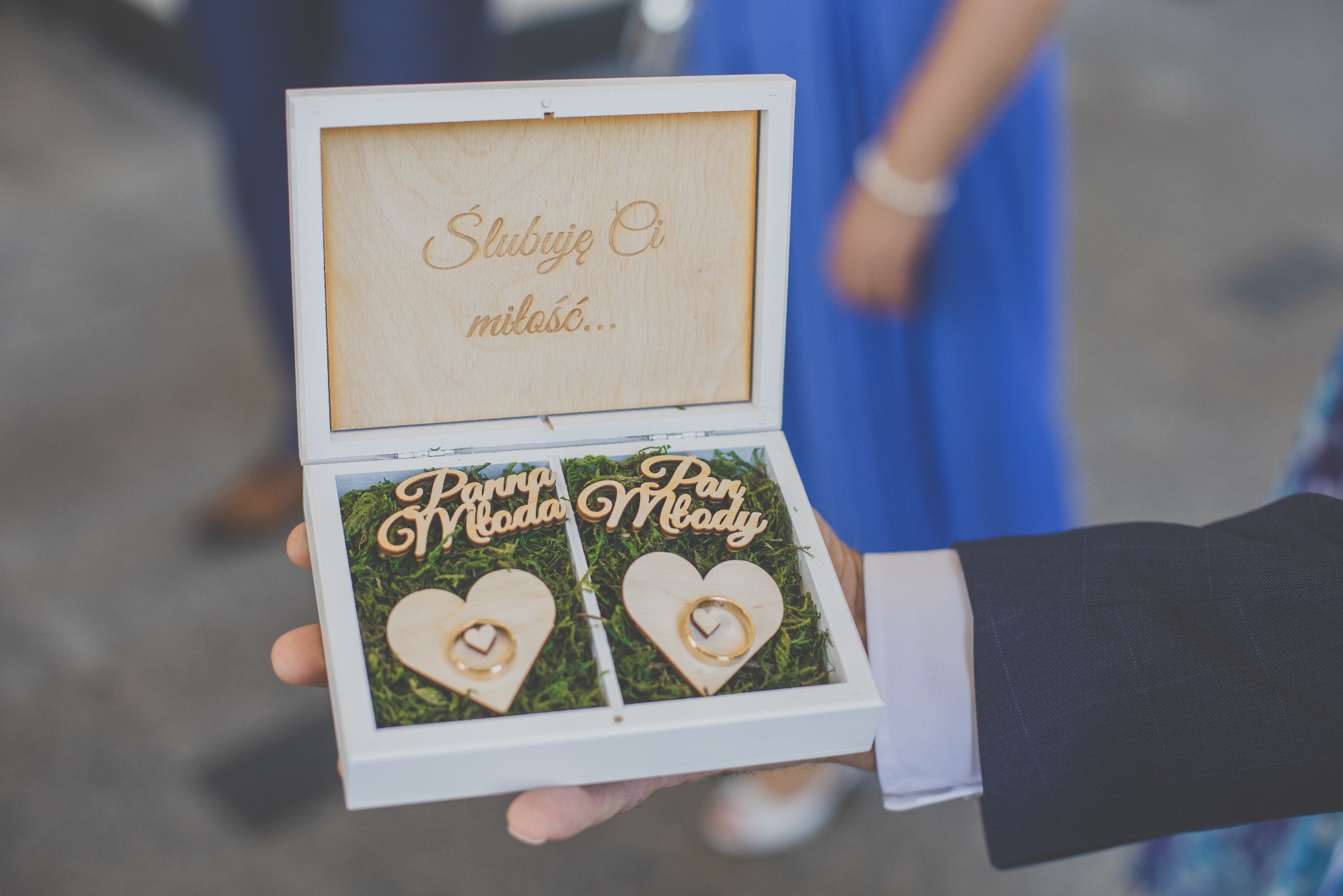 Otwarta, biała szkatułka na obrączki z napisem 'Ślubuję Ci miłość...', wypełniona mchem i drewnianymi sercami z obrączkami, trzymana w dłoni mężczyzny w garniturze.
