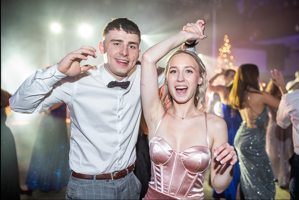 Uśmiechnięta para w tańcu na studniówce, on w białej koszuli i muszce, ona w różowej sukience, w tle rozświetlona choinka i bawiący się ludzie.