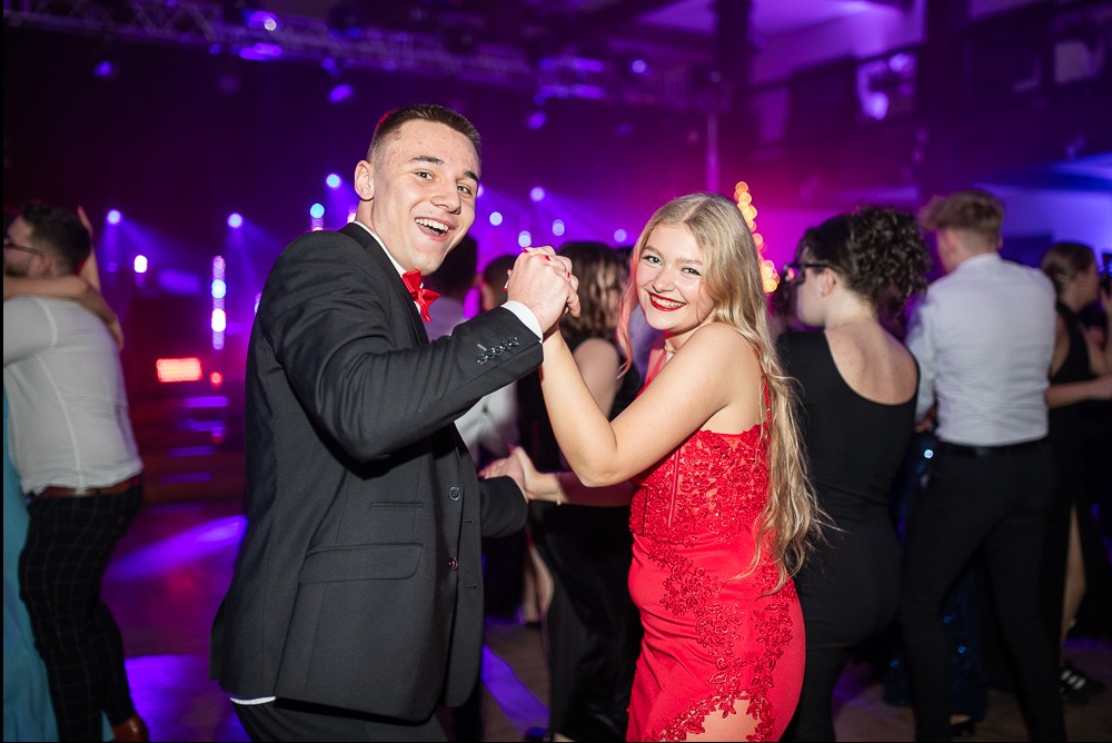 Uśmiechnięta para na studniówce, on w czarnym garniturze z czerwoną muchą, ona w czerwonej sukience, tańczą na tle rozświetlonej sali balowej.