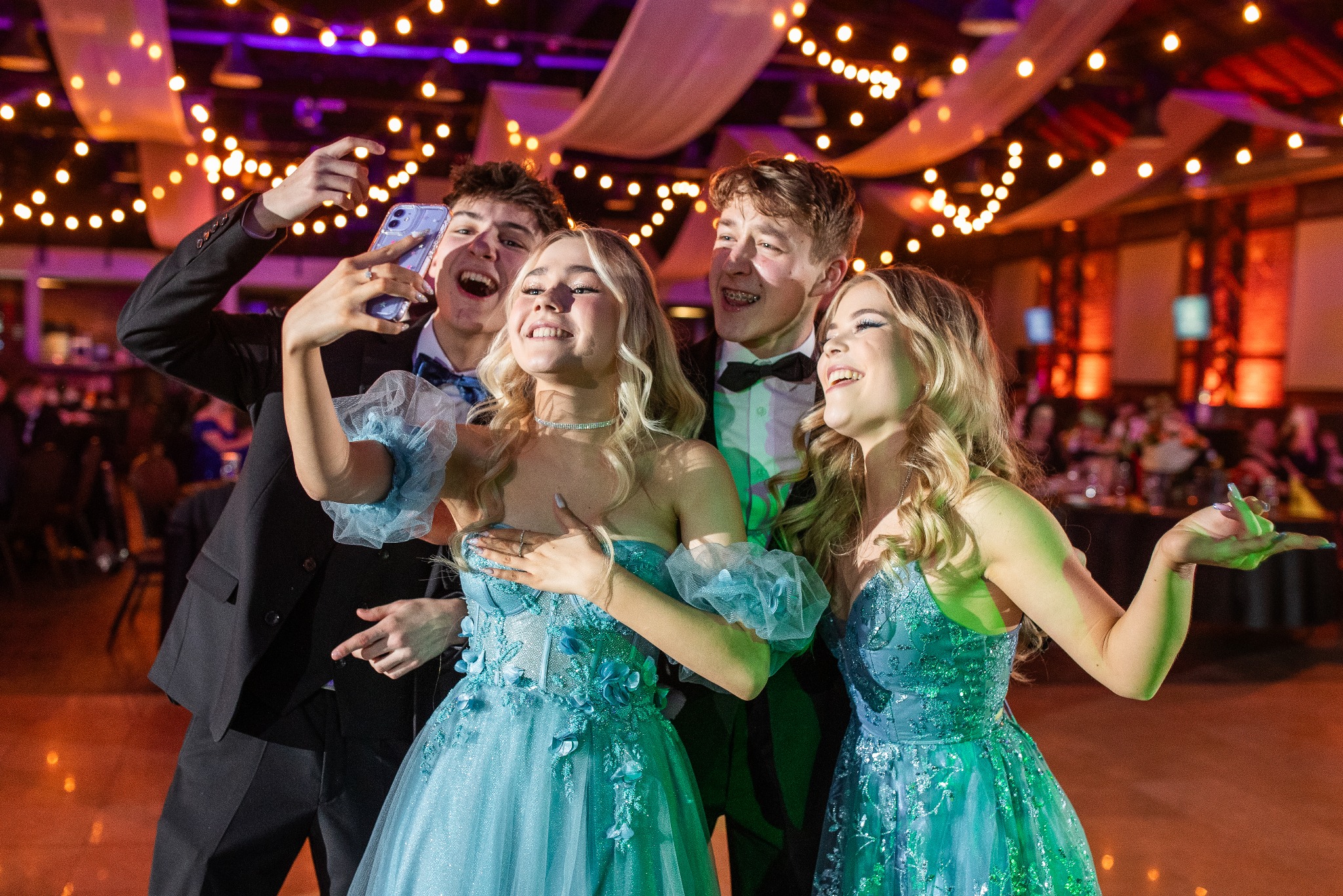 Uśmiechnięta grupa nastolatków w strojach wieczorowych robi selfie na tle dekoracji sali balowej z oświetleniem punktowym i drapowanymi tkaninami.