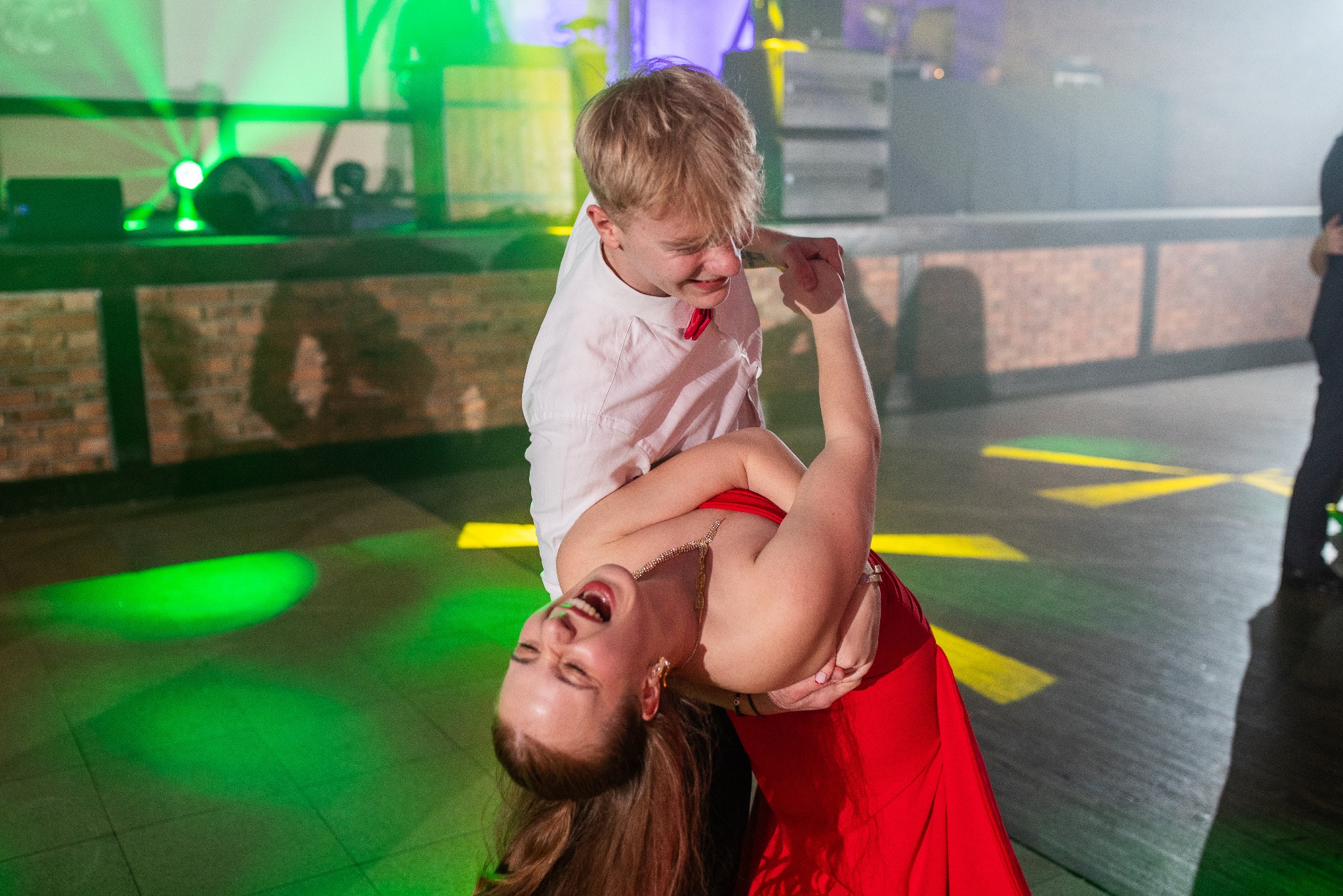 Studniówka w obiektywie: dynamiczne ujęcie tańczącej pary, dziewczyna w czerwonej sukni, chłopak w białej koszuli, parkiet oświetlony zielonym światłem, radosne emocje.