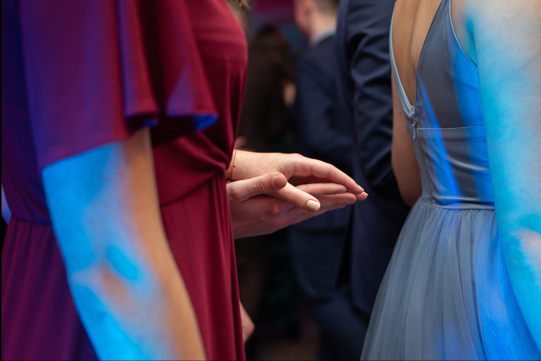 Dłonie dwóch osób w sukienkach (bordowej i błękitnej) na tle innych uczestników wydarzenia, oświetlone niebieskim światłem. Widoczne detale sukienek i subtelny manicure.
